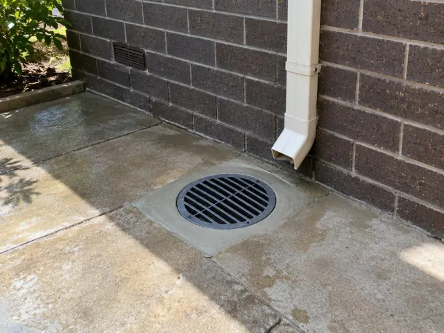 Outdoor drain grate beside brick wall and downpipe on paved patio in Birmingham Gardens
